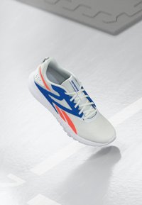 Weiße Sportschuh mit blauen und roten Akzenten, geschnürt und schwebend über einem hellgrauen Boden mit einer Linie und Matte im Hintergrund.