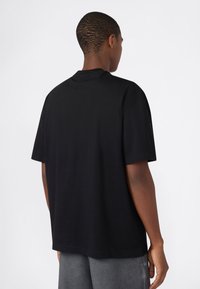 Persona girata di spalle che indossa una semplice maglietta nera a maniche corte e pantaloni grigi su uno sfondo chiaro e neutro.