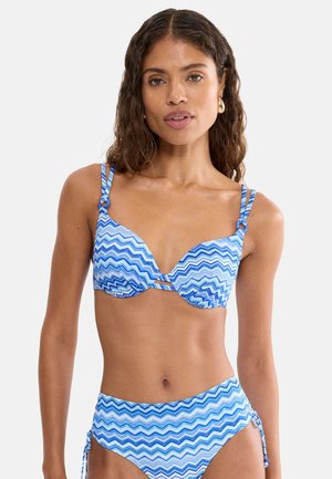 Frau mit welligem dunklem Haar, die einen blau-weiß gezackten Bikini mit hoch taillierten Hosen und goldene Creolen auf weißem Hintergrund trägt.