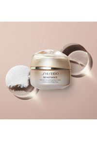 Shiseido Benefiance Wrinkle Smoothing Eye Cream i en rund, beige burk med en gulddetalj och ett glansigt lock, tillsammans med en cirkulär gel och puder.