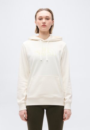 Jonge vrouw die een crèmekleurige hoodie draagt met een voorzak en een vage gele "The North Face"-logo, gecombineerd met donkerolijfgroene broek.