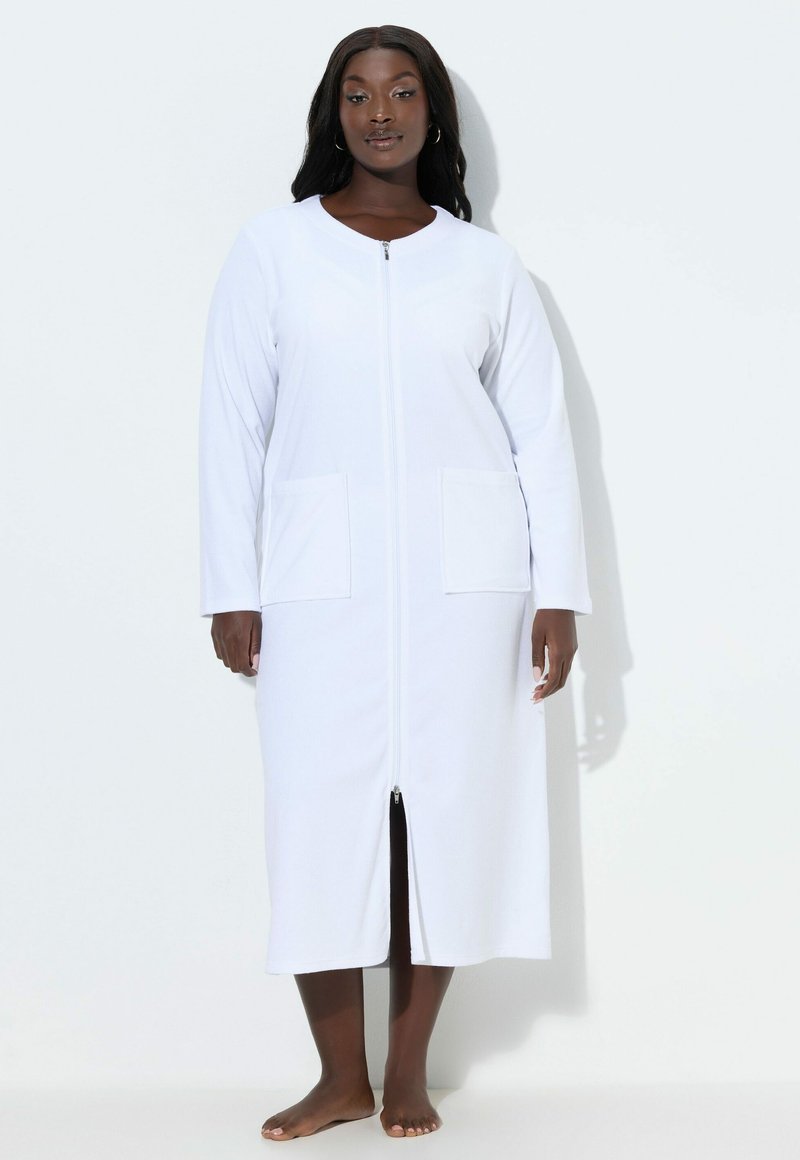 Schwarze Frau steht barfuß und trägt ein langes weißes Kleid mit Reißverschluss und Taschen vorne vor einem einfarbigen Hintergrund.