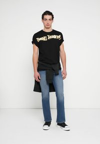 Μαύρο βαμβακερό T-shirt με χρυσό γραφικό κείμενο, συνδυασμένο με μπλε τζιν παντελόνι με ίσια γραμμή και μαύρα αθλητικά παπούτσια, με μαύρο φούτερ δεμένο στη μέση.