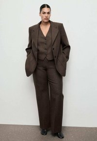 Completo marrone su misura con blazer oversize, gilet con bottoni e pantaloni a gamba larga. Realizzato in tessuto testurizzato, con eleganti calzature nere.