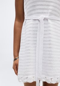 Πλεκτό λευκό φόρεμα με ζώνη δεμένη στη μέση, φορεμένο από ένα άτομο με ασημένιο βραχιόλι στον αριστερό καρπό.