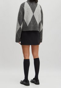 Personne portant un pull à losanges gris, une mini-jupe noire, des chaussettes noires montantes jusqu'aux genoux, et des mocassins noirs, debout devant un fond uni clair.