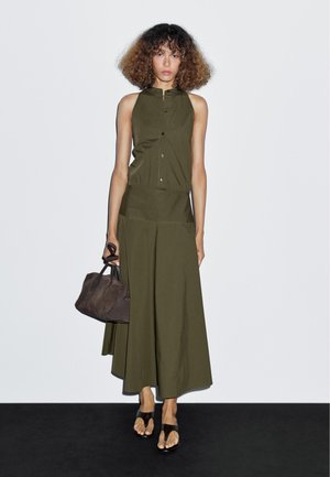 Abito senza maniche verde oliva con bottoni frontali e vita aderente, abbinato a una borsa marrone scuro e sandali neri. Texture liscia.