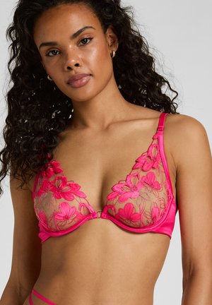 Frau trägt hellrosa floralen Spitzen-BH mit verstellbaren Trägern, zeigt Oberkörper und Gesicht vor neutralem Hintergrund.