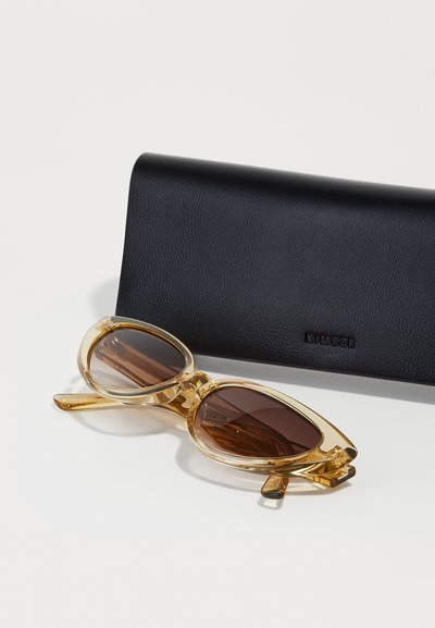 Gafas de sol amarillas claras con lentes de tono marrón, en forma ovalada y con montura angular. Estuche negro texturizado con el logo "KIMEZE" en relieve.