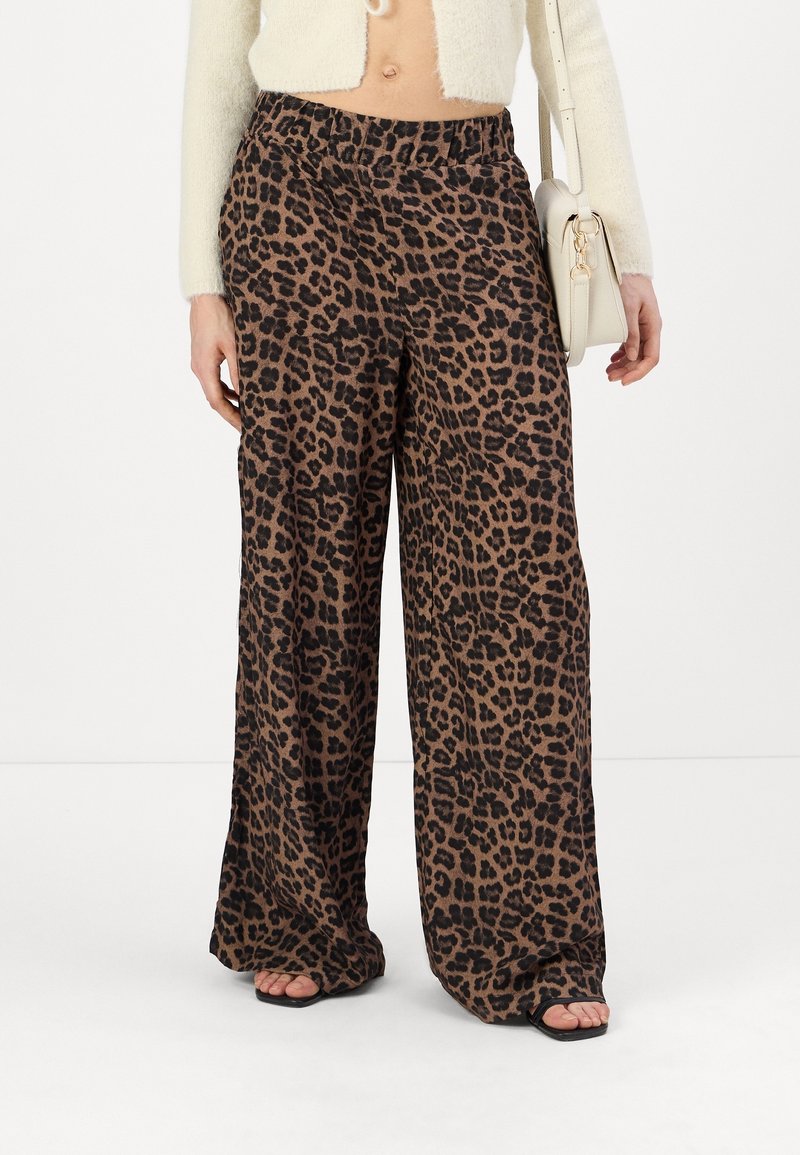 Pantalons larges en tissu à imprimé léopard marron et noir avec une taille élastique, associés à des sandales noires à talons ouverts et un sac à main beige.