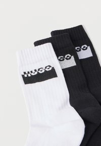 Tres pares de calcetines tipo tripulación: uno blanco, uno negro y uno gris. Cada par presenta una banda de logo en contraste con "HUGO" en fuente negrita.