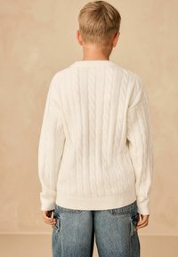 Maglione bianco in maglia a trecce con polsini e orlo a coste, caratterizzato da una vestibilità ampia. Indossato sopra un paio di jeans baggy azzurri su uno sfondo neutro.