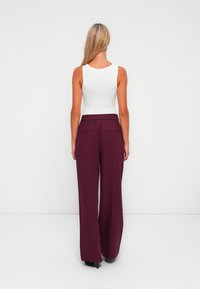 Donna con lunghi capelli biondi, che indossa una canotta bianca senza maniche e pantaloni larghi viola, in piedi davanti a uno sfondo bianco uniforme.