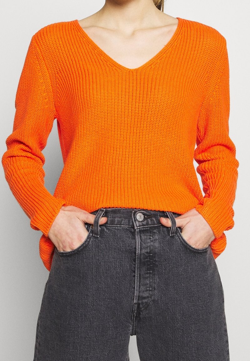 Helles orange, geripptes Strickpullover mit V-Ausschnitt und langen Ärmeln, kombiniert mit hochgeschnittenen dunkelgrauen Jeans. Strukturiertes Material.