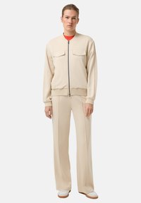 Béžová bomber bunda na zip se dvěma kapsami na hrudi, doplněná širokými kalhotami. Hladká textura, neformální design a žebrované manžety.