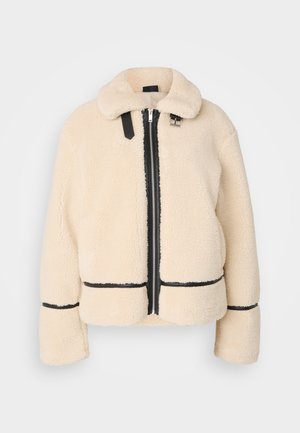 Helle, flauschige Teddyjacke in Cremefarbe mit einem Reißverschluss vorne, schwarzem Reißverschluss und Lederakzenten. Sie hat einen weichen, kragenartigen Hals und weite Ärmel.