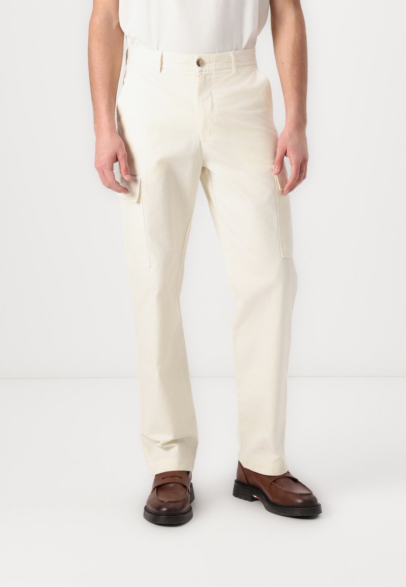 Pantalons cargo en tissu blanc cassé, présentant des poches latérales, une coupe droite et une fermeture à bouton à la taille. Portés avec des chaussures marron foncé.