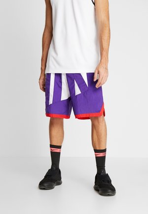 Mężczyzna w białym podkoszulku bez rękawów, fioletowo-czerwonych spodenkach do koszykówki, czarnych trampkach i czarnych skarpetkach z różowym logo Nike stoi na białej podłodze.