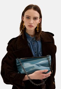 Tyrkysová lakovaná kabelka s řetízkem, která se vyznačuje hladkým povrchem a jemně zmačkaným designem. Kovové detaily na vrchu.