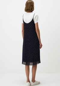 Navyblauwe midi-jurk met een subtiel kruispatroon, schouderbanden en een doorschijnende zoom. Gecombineerd met een wit T-shirt en witte sneakers.