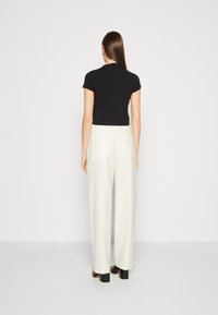 Schwarzes Kurzarmoberteil kombiniert mit cremefarbenen weit geschnittenen Hosen, die zwei Gesäßtaschen und eine taillierte Taille aufweisen, vor einem neutralen Hintergrund.