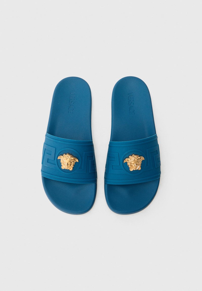 Blaue Gummisandalen mit strukturierter Oberfläche, versehen mit einem goldenen Medusa-Emblem und einem geometrischen Muster auf dem Riemen.