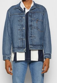 Blå denimjacka med en klassisk passform, knäppning, bröstficka och en något urtvättad textur. Bärs över en marinblå och vit randig skjorta.