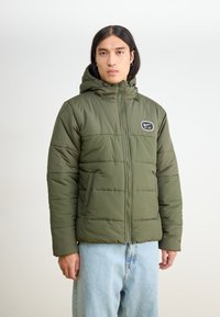 Alyvuogių žalios spalvos pūkinė striukė su užtrauktuku, su gaubtu ir logotipo aplikacija ant krūtinės. Turi siūtuotą dizainą ir šonines kišenes.