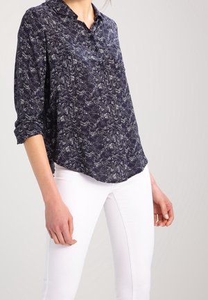 Navy blue blouse in a lightweight fabric with a subtle abstract pattern, featuring a collar and rolled sleeves, paired with white pants.