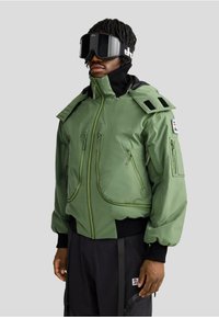 Veste isolante verte avec plusieurs poches zippées, col incliné et accents noirs. Portée avec de grandes lunettes de ski noires.