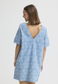 Femme aux cheveux mi-longs portant une robe bleu clair à manches courtes avec des motifs floraux blancs et un dos en V profond, souriante et regardant sur le côté.