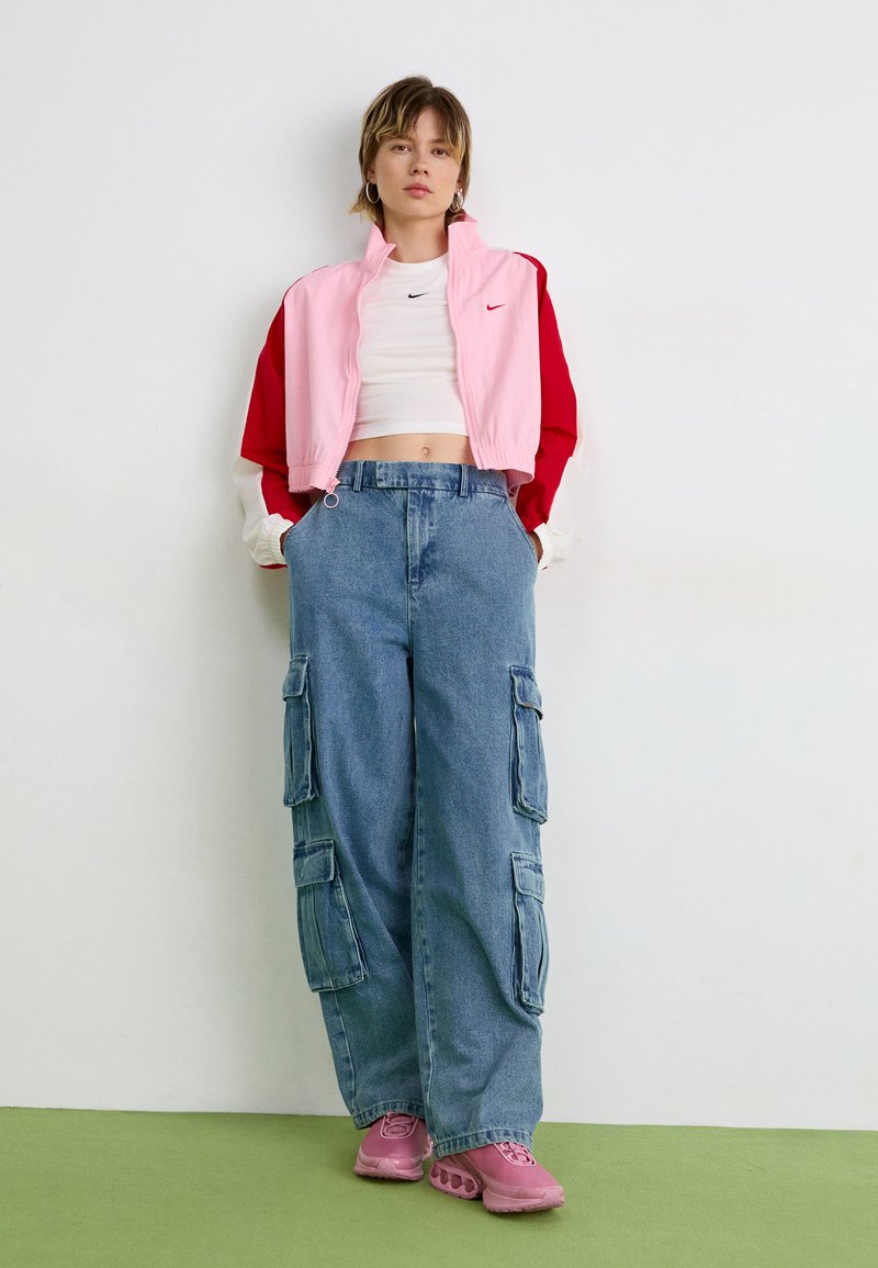 Zuschneidete pinke und rote Jacke über einem weißen Oberteil, kombiniert mit hellblauen Cargo-Hosen und pinken Sneakers, vor einem weißen Hintergrund stehend.