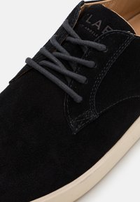 Schwarze Wildleder-Schnürschuhe mit grauen Schnürsenkeln, ausgestattet mit einer hellen Sohle und dezenten Stickdetails nahe dem oberen Rand.