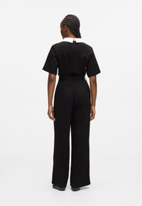 Čierny overal s krátkymi rukávmi a bielym golierom. Má priliehavý pás a široký strih nohavíc, hladkú textúru a zapínanie na zips na chrbte.