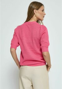 Rosa gestrickter Pullover mit kurzen, puffartigen Ärmeln; strukturiertes Muster und schmaler, gerippter Saum. Kombiniert mit beigen Hosen.