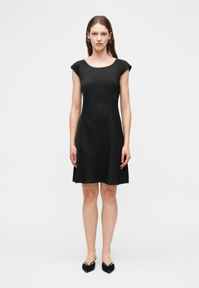 Γυναίκα που στέκεται με το σώμα προς τα εμπρός, φορώντας μαύρο φόρεμα μέχρι το γόνατο με κοντά μανίκια και μαύρα παπούτσια με μυτερό μπροστινό μέρος, σε απλό φόντο.