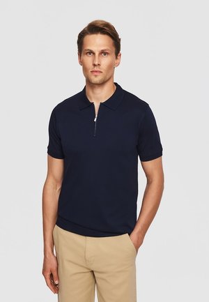 Mężczyzna o jasnej karnacji i krótkich brązowych włosach, ubrany w granatową koszulkę polo z krótkim rękawem i zamkiem błyskawicznym oraz beżowe spodnie, stojący na jednolitym tle.