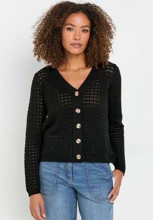 Cardigan noir en tricot avec un col en V, six boutons en bois, des manches longues et une texture en maille ouverte. Associé à un jeans bleu clair.