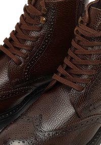 Brązowe skórzane botki za kostkę o strukturalnym wykończeniu, z okrągłym noskiem i zdobieniami w stylu brogue. Wyposażone w brązowe sznurowadła i mosiężne oczka.