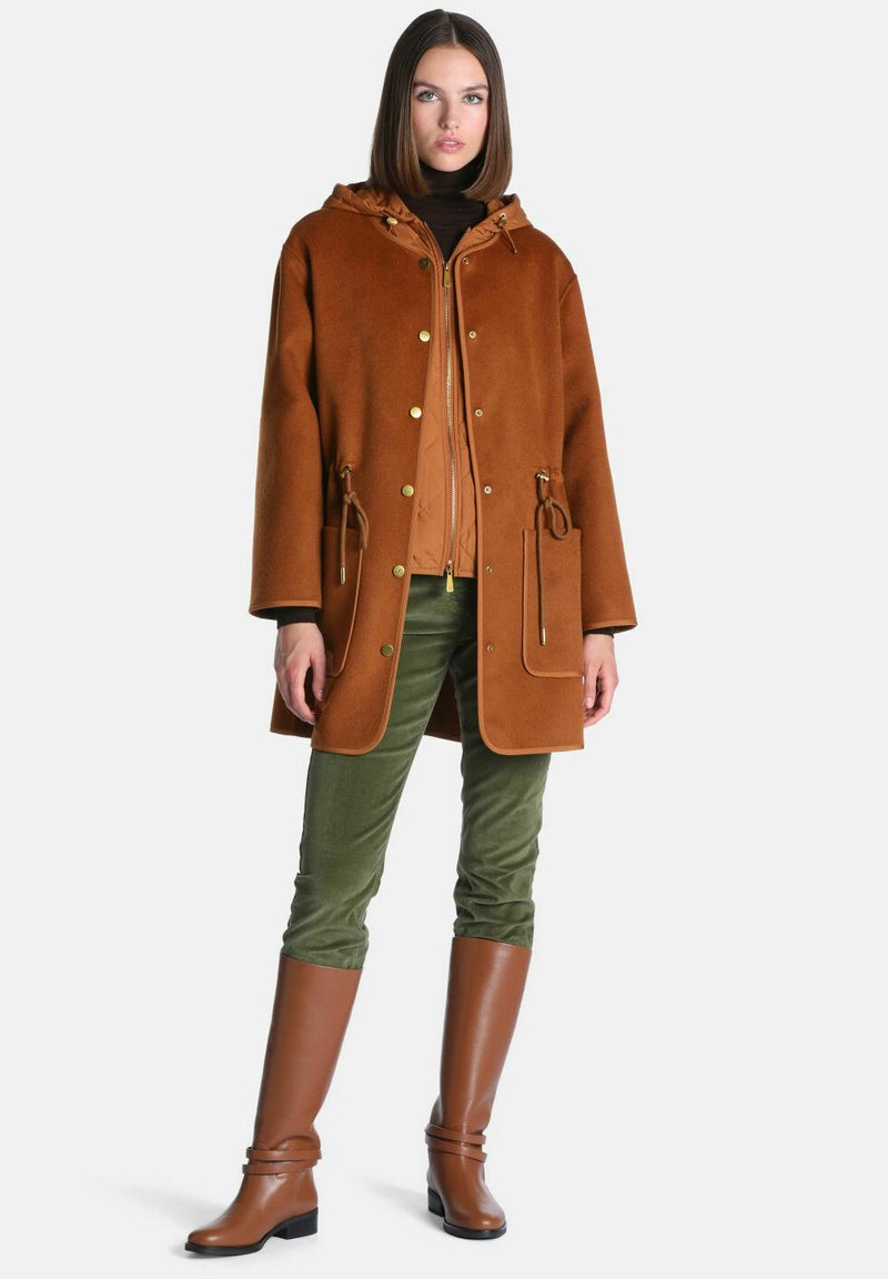 Femme portant un manteau marron à capuche avec poches avant, pantalon vert et bottes en cuir marron montant jusqu'aux genoux, debout sur un fond uni.