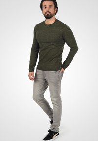 Donker groene trui met lange mouwen en een textuurgebreid patroon, gecombineerd met lichtgrijze jeans en zwarte schoenen met gouden accenten.