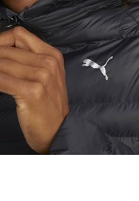 Chaqueta de plumas negra con cuello alto, cierre de cremallera y un logo de Puma plateado. El material tiene un aspecto suave y ligeramente brillante, con secciones acolchadas.