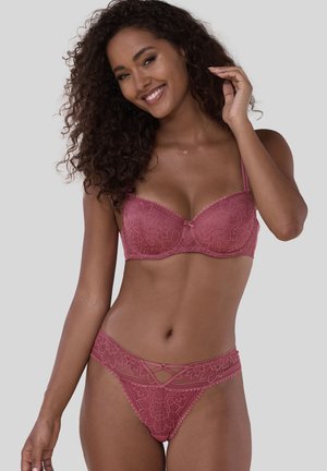Woman with curly hair smiling, wearing matching pink lace bra and panties with floral patterns and delicate detailing.