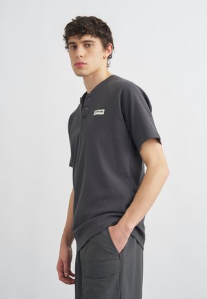 Mladi moški s kodrastimi temnimi lasmi, oblečen v temnosivo majico Henley s kratkimi rokavi in temne hlače, z eno roko v žepu.