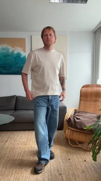 Camisa de manga corta beige, jeans azules, zapatos negros. De pie en una habitación con un sofá gris, una silla de mimbre y arte mural decorativo.