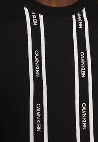Černé bavlněné tričko s vertikálními bílými pruhy, na kterých je vytištěn text "CALVIN KLEIN". Kulatý výstřih a hladká textura.