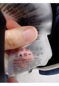 Etiqueta de cuidado sostenida por una mano, con varios símbolos de cuidado en negro sobre fondo blanco. El texto aconseja lavar a un máximo de 30°C, proceso suave, sin blanqueo.