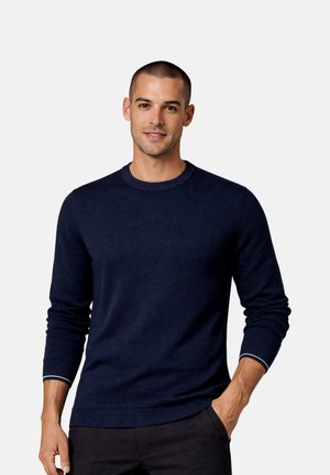 Man with short hair wearing a navy blue long-sleeve sweater and dark pants, standing with one hand in pocket against a plain background.