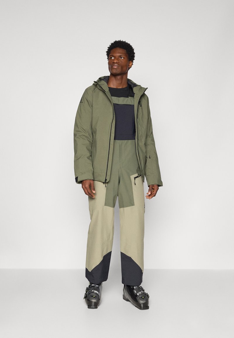 Wasserabweisende Jacke in Olivgrün, kombiniert mit farblich abgestimmten Hosen in hellbeige und dunkelblau. Verfügt über eine Kapuze und ein verstellbares Design.