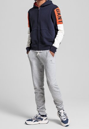 Mężczyzna w granatowej bluzie z kapturem zapinanej na zamek, z pomarańczowymi i białymi rękawami, szarych spodniach dresowych i niebiesko-białych butach sportowych, stojący z rękami w kieszeniach.